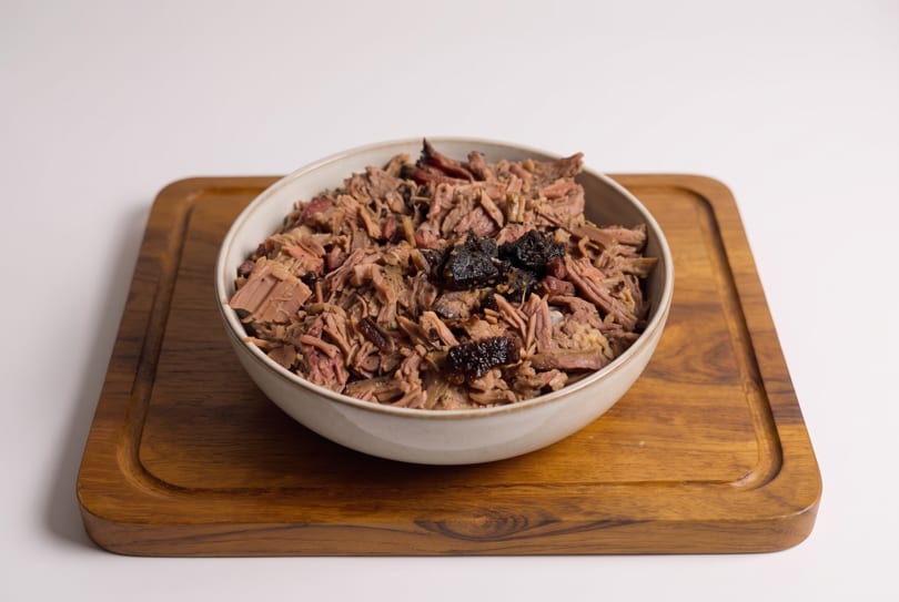 Shredded meat with dark pieces, possibly prunes, in a white bowl on a wooden cutting board.