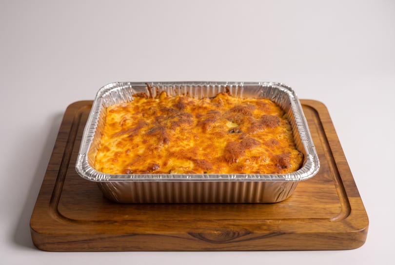 Baked pasta with melted cheese in a foil pan on a wooden cutting board.