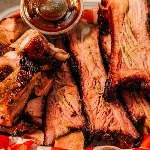 Close-up of smoked ribs and brisket in a container with a side of coleslaw and BBQ sauce.