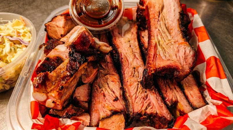 Close-up of smoked ribs and brisket in a container with a side of coleslaw and BBQ sauce.