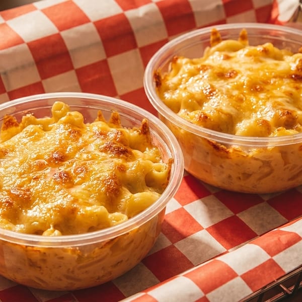 Two clear plastic bowls of baked mac and cheese with a crispy topping, on red and white checkered paper.