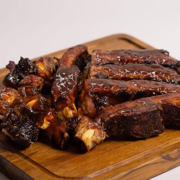 Glazed BBQ ribs piled on a wooden cutting board against a white background.