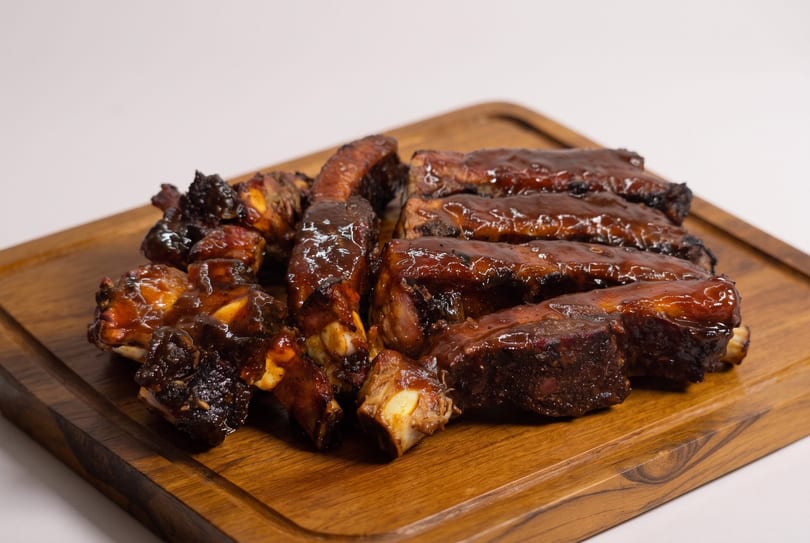 Glazed BBQ ribs piled on a wooden cutting board against a white background.