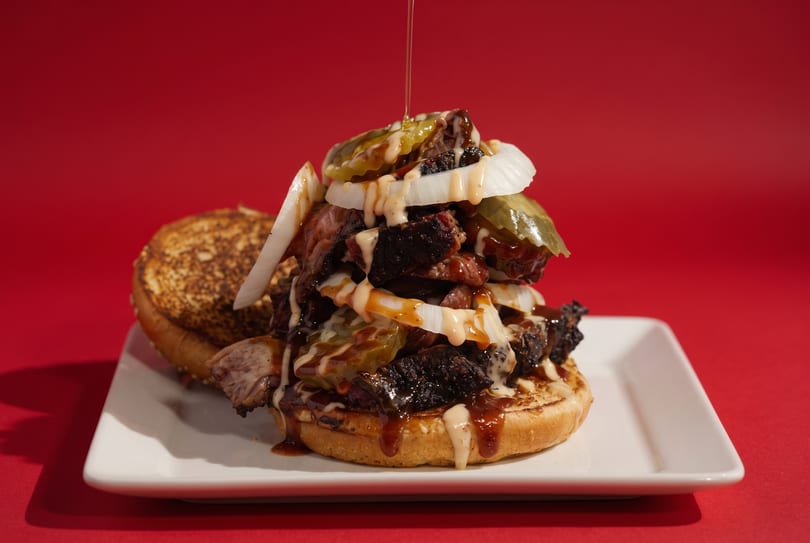 Large BBQ sandwich with brisket, onions, pickles, and sauce drizzling on a white plate.
