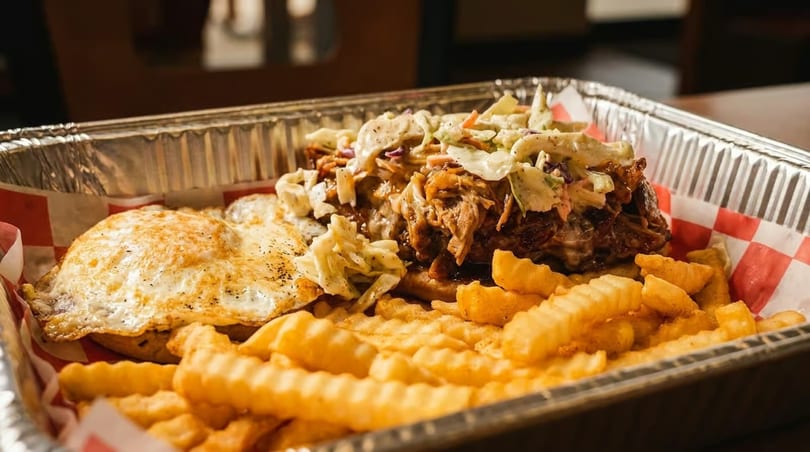 Pulled pork sandwich with coleslaw, a fried egg, and crinkle-cut fries in a foil tray.