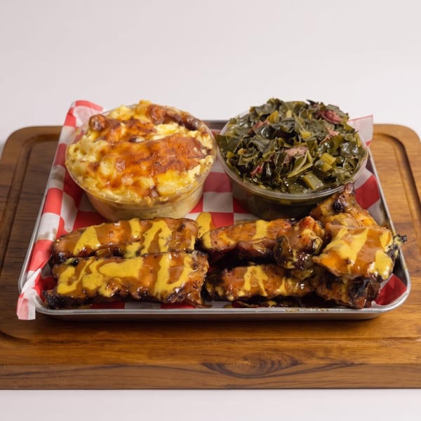 A tray of BBQ ribs with mustard sauce, mac and cheese, and collard greens on a wooden board.