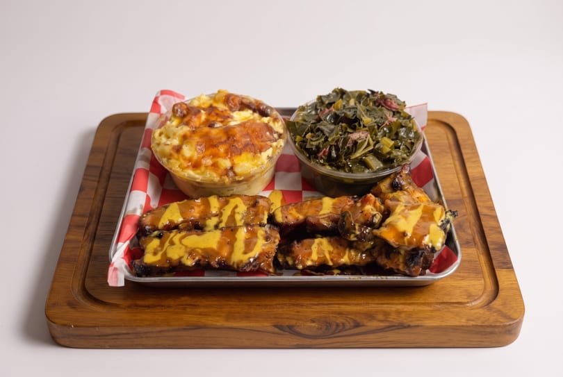 A tray of BBQ ribs with mustard sauce, mac and cheese, and collard greens on a wooden board.