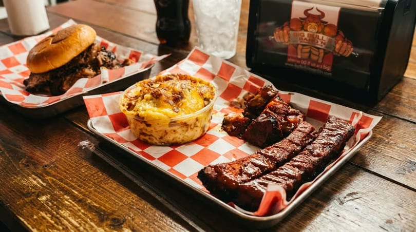 Big Bull Bar-B-Q'N ribs, mac and cheese, and a pulled pork sandwich on a wooden table.